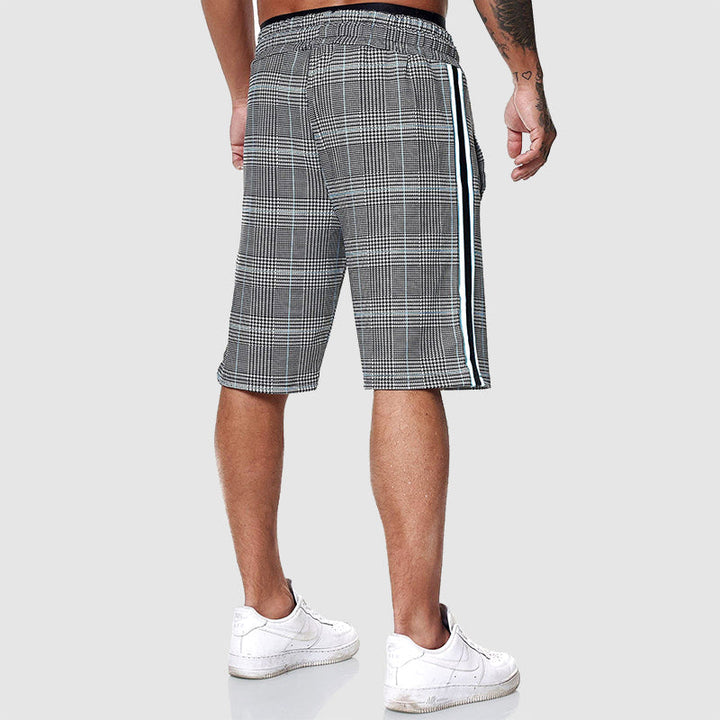 Shorts uomo a quadri grigi con strisce laterali bianche, indossati con scarpe da ginnastica bianche. Stile casual, moda estiva.
