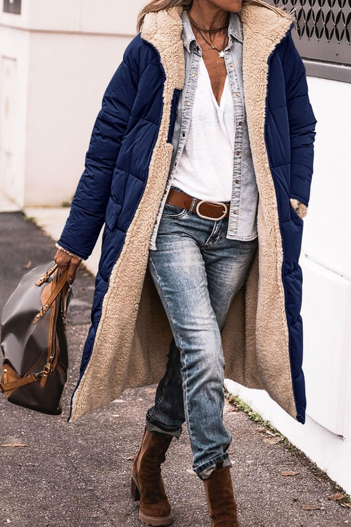 Donna con cappotto lungo blu imbottito, camicia bianca, jeans, stivali marroni e borsa in pelle. Moda invernale casual chic.