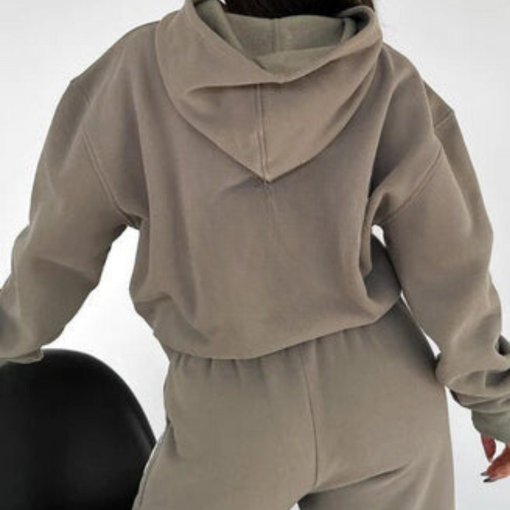 Donna con felpa con cappuccio beige, pantaloni abbinati, seduta su sedia nera. Abbigliamento casual, moda autunnale, stile confortevole.