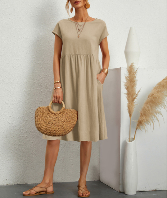 Donna con abito beige estivo, borsa di paglia intrecciata, sandali marroni, accessori dorati, accanto a vaso bianco con pampas. Moda casual elegante.