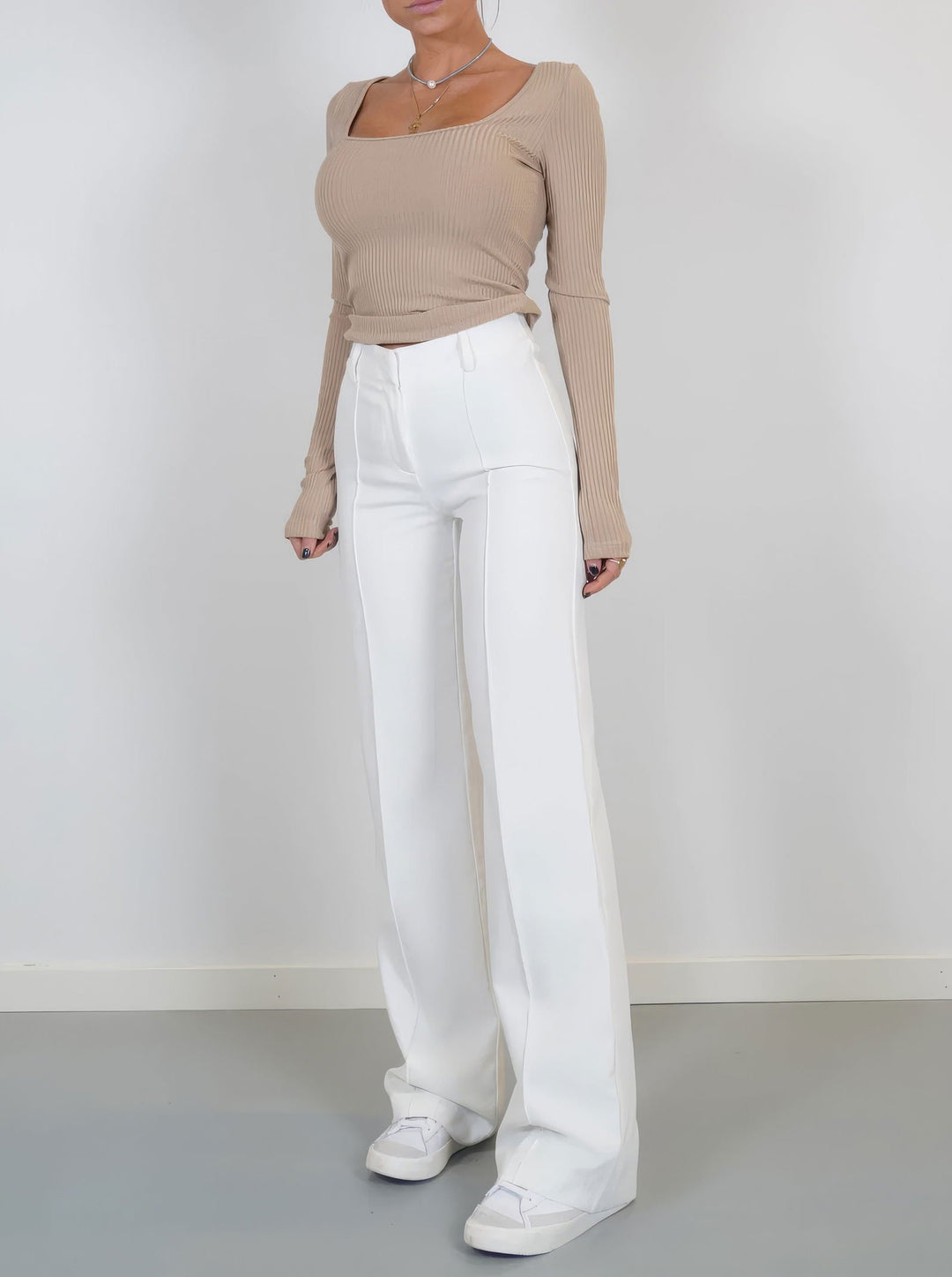 Donna in maglia beige a coste e pantaloni bianchi a vita alta, scarpe da ginnastica bianche. Moda casual elegante, abbigliamento femminile trendy.