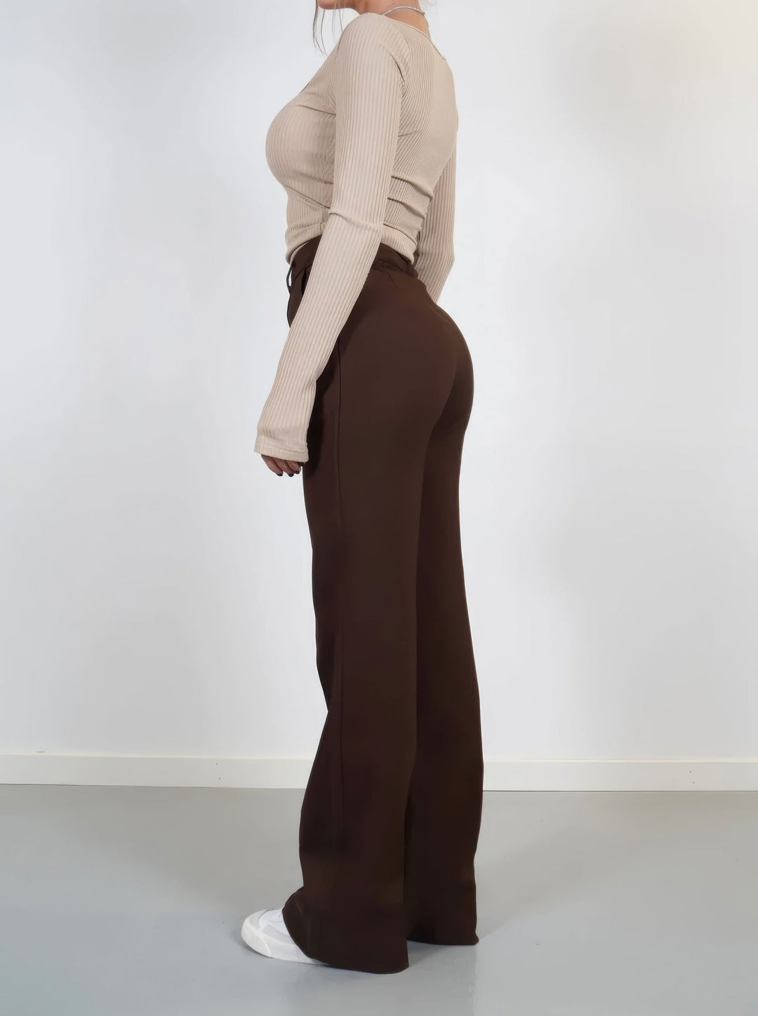 Donna in pantaloni a zampa marroni e maglia beige a coste, vista laterale. Moda autunnale, abbigliamento casual elegante, scarpe bianche.
