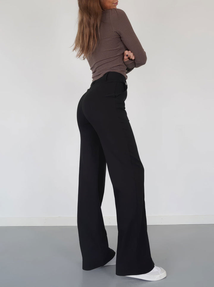 Donna in pantaloni neri a vita alta, maglia marrone a maniche lunghe, scarpe bianche; moda casual elegante, abbigliamento femminile autunnale.