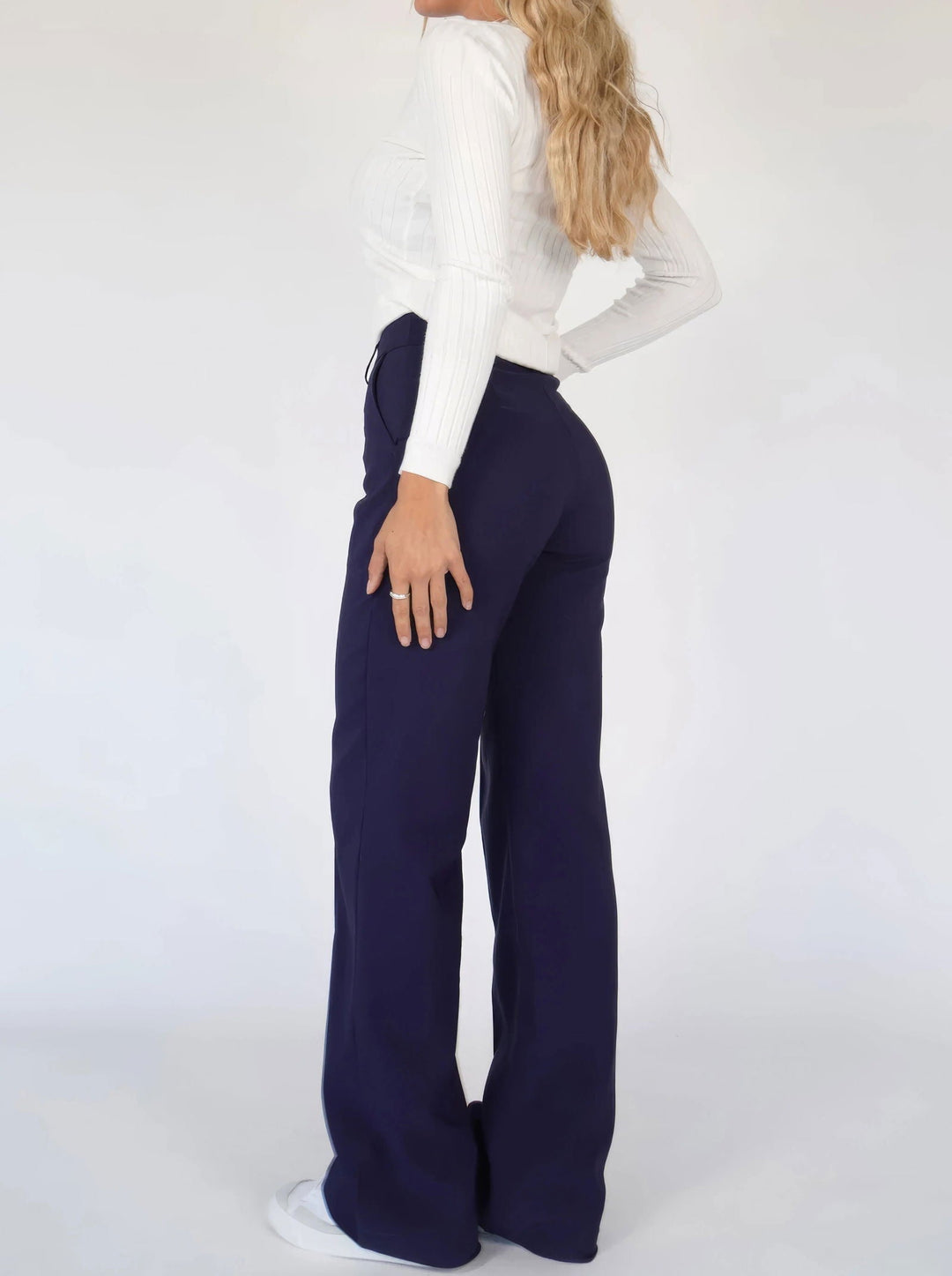 Donna in pantaloni blu scuro a vita alta, maglia bianca a maniche lunghe, posa laterale. Moda femminile, abbigliamento casual elegante.