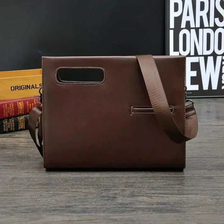 Borsa a tracolla in pelle marrone scuro, design elegante e moderno, con manico integrato e cinturino regolabile, su sfondo di libri e poster.