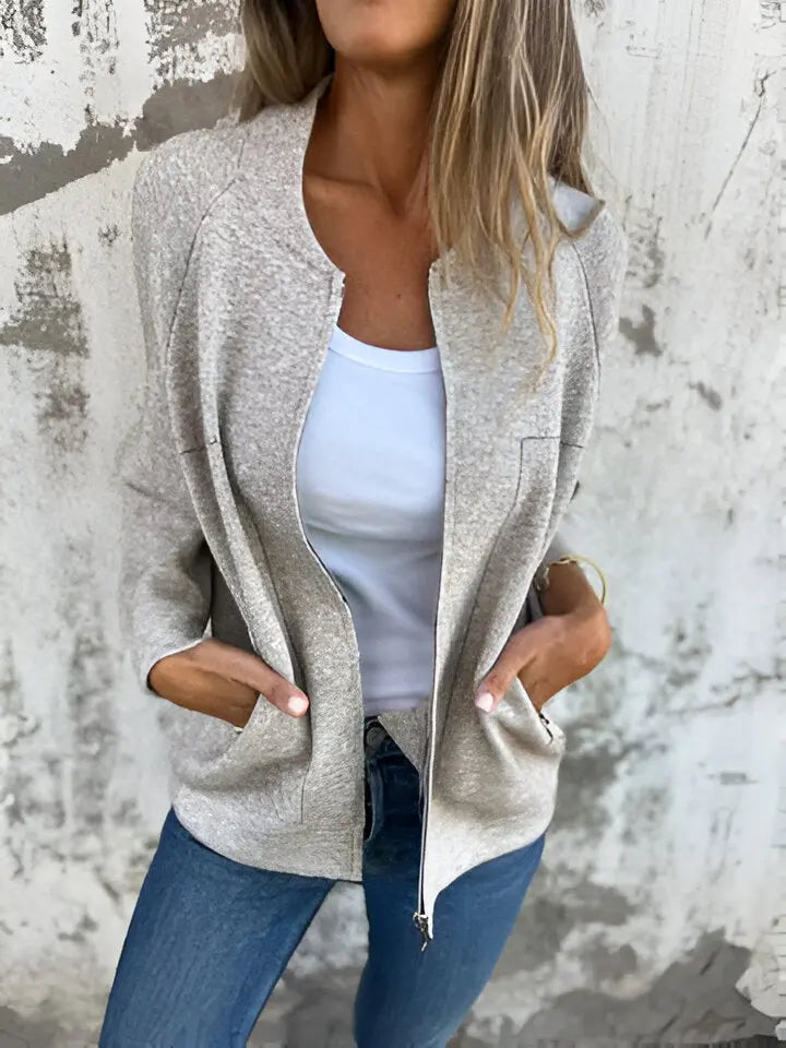 Donna con giacca beige aperta, top bianco e jeans blu davanti a muro grigio. Moda casual, abbigliamento femminile, stile urbano.