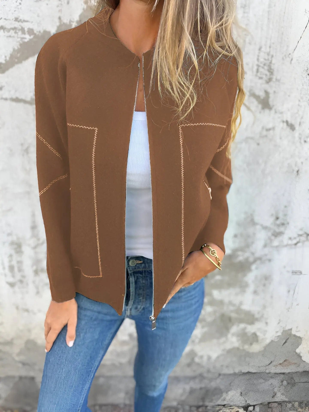 Giacca marrone donna, zip aperta, su top bianco e jeans blu. Moda casual autunnale, stile elegante. Sfondo muro grigio.