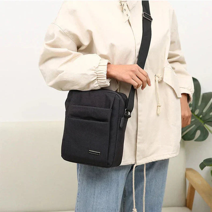 Borsa a tracolla nera unisex, design minimalista, indossata su giacca beige e jeans. Ideale per moda casual e praticità quotidiana.