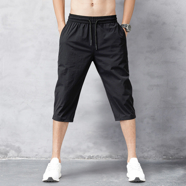Pantaloni corti sportivi neri da uomo, lunghezza al polpaccio, con elastico in vita e tasche laterali, indossati con scarpe da ginnastica bianche.