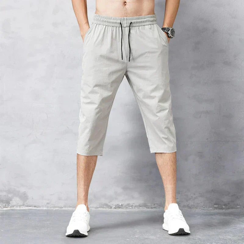 Pantaloni corti sportivi da uomo grigi, lunghezza al polpaccio, con elastico in vita e coulisse, abbinati a scarpe da ginnastica bianche.