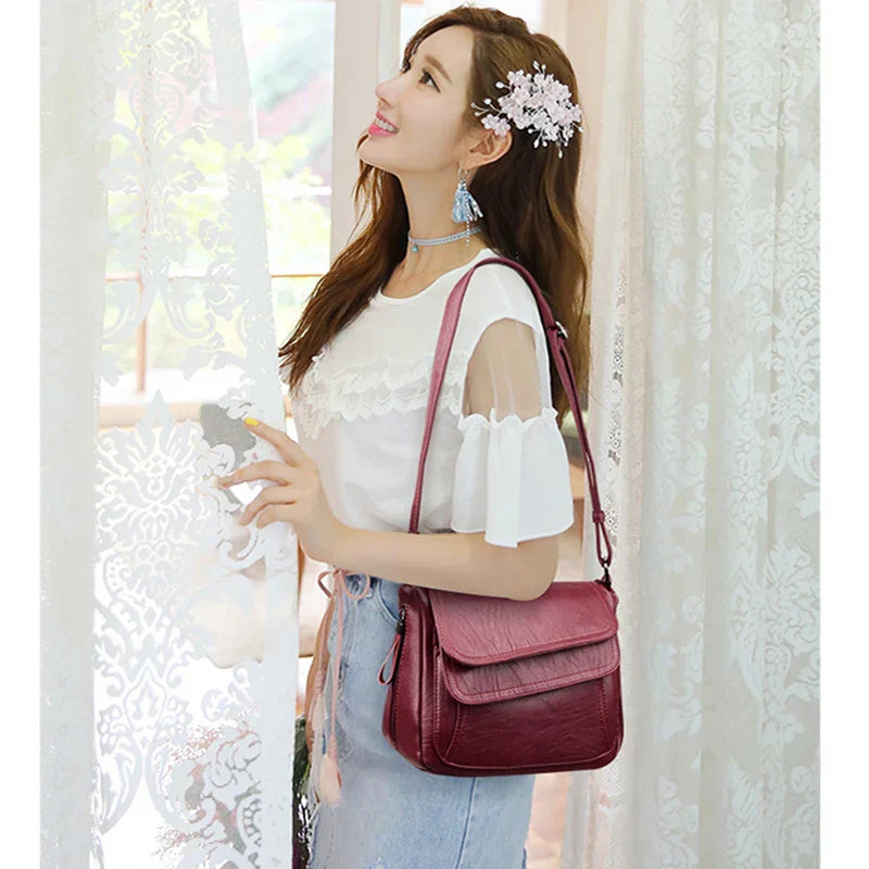 Donna sorridente con borsa a tracolla rossa in pelle, indossa abito bianco estivo, accessori floreali nei capelli, vicino a finestra con tenda.