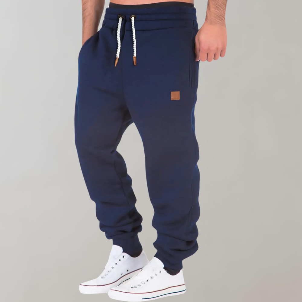 Pantaloni tuta blu uomo, cordoncino bianco, tasche laterali, etichetta marrone, abbinati a scarpe da ginnastica bianche.