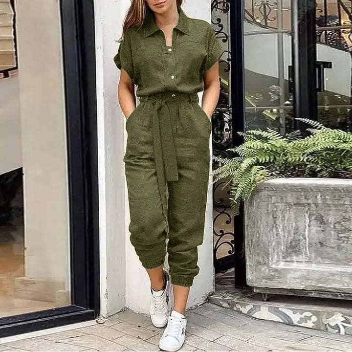 Donna con tuta verde oliva, maniche corte, cintura in vita, davanti a una vetrina. Indossa scarpe da ginnastica bianche. Moda casual elegante.