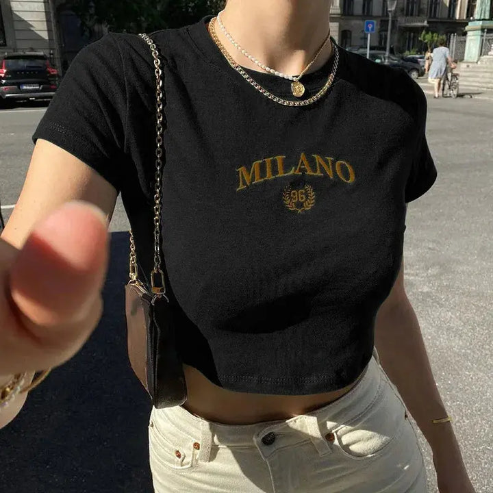 Donna con t-shirt nera "Milano", catena dorata, borsa a tracolla marrone, jeans chiari. Moda urbana estiva, stile casual chic.