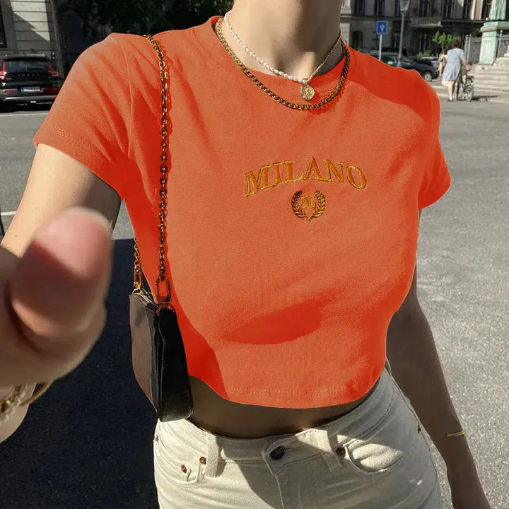 Donna con t-shirt arancione "Milano", collana dorata, borsa a tracolla marrone, jeans chiari. Moda estiva urbana, street style italiano.