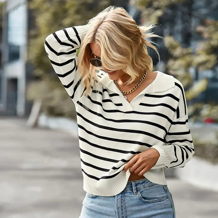 Maglione a righe bianche e nere, donna bionda, occhiali da sole, collana dorata, jeans chiari, moda casual autunnale, sfondo urbano sfocato.