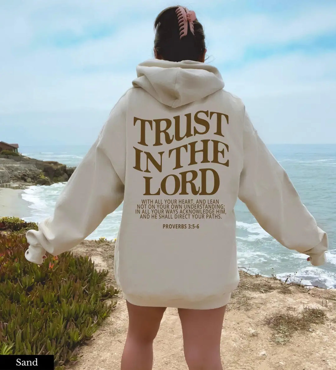 Donna in felpa beige con scritta "Trust in the Lord", vista mare. Abbigliamento casual, moda ispirata alla fede, paesaggio costiero.