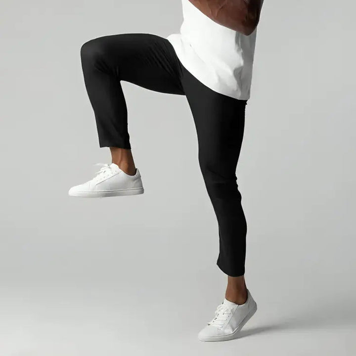 Pantaloni sportivi neri da uomo, elasticizzati, indossati con scarpe da ginnastica bianche e maglietta bianca. Ideali per fitness e allenamento.