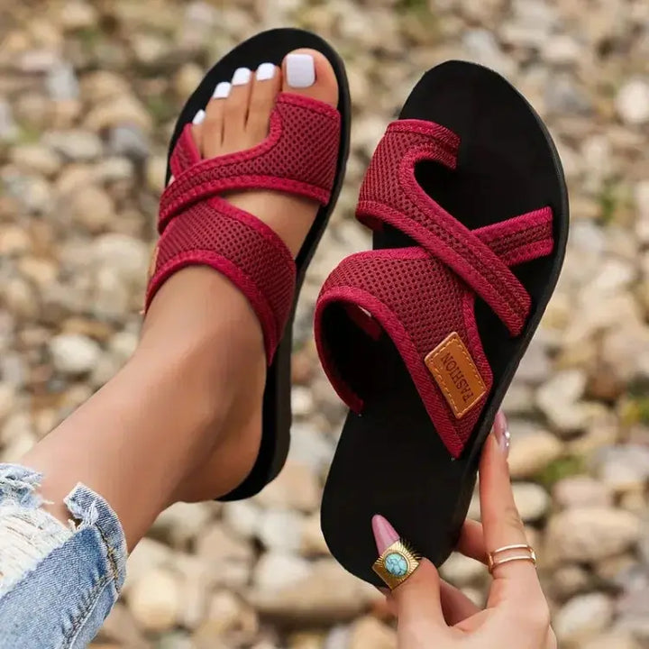 Sandali rossi da donna con suola nera, design intrecciato, indossati su sfondo di ghiaia. Scarpe estive eleganti e comode.