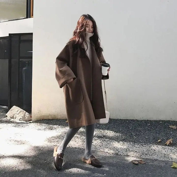 Donna con cappotto marrone oversize, maglione a collo alto, leggings grigi e mocassini marroni, tiene una tazza da caffè. Moda autunnale casual.