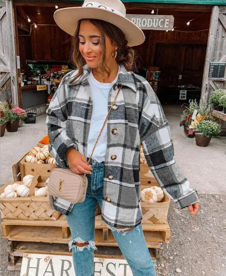 Donna con cappotto a quadri grigio e bianco, jeans strappati e borsa beige, davanti a un mercato agricolo con zucche e piante. Moda autunnale casual.