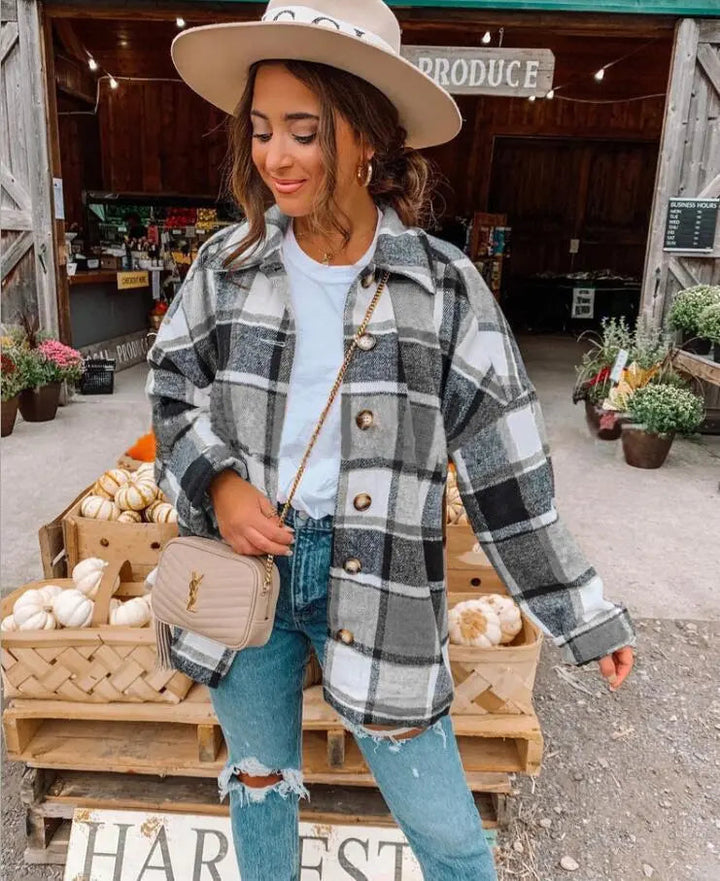 Donna con cappotto a quadri grigio e bianco, jeans strappati e borsa beige, davanti a un mercato agricolo con zucche e piante. Moda autunnale casual.