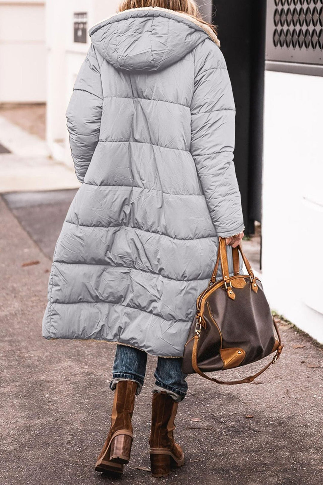 Donna con cappotto lungo imbottito grigio, borsa in pelle marrone, stivali invernali, strada urbana. Moda invernale, abbigliamento caldo, stile casual.