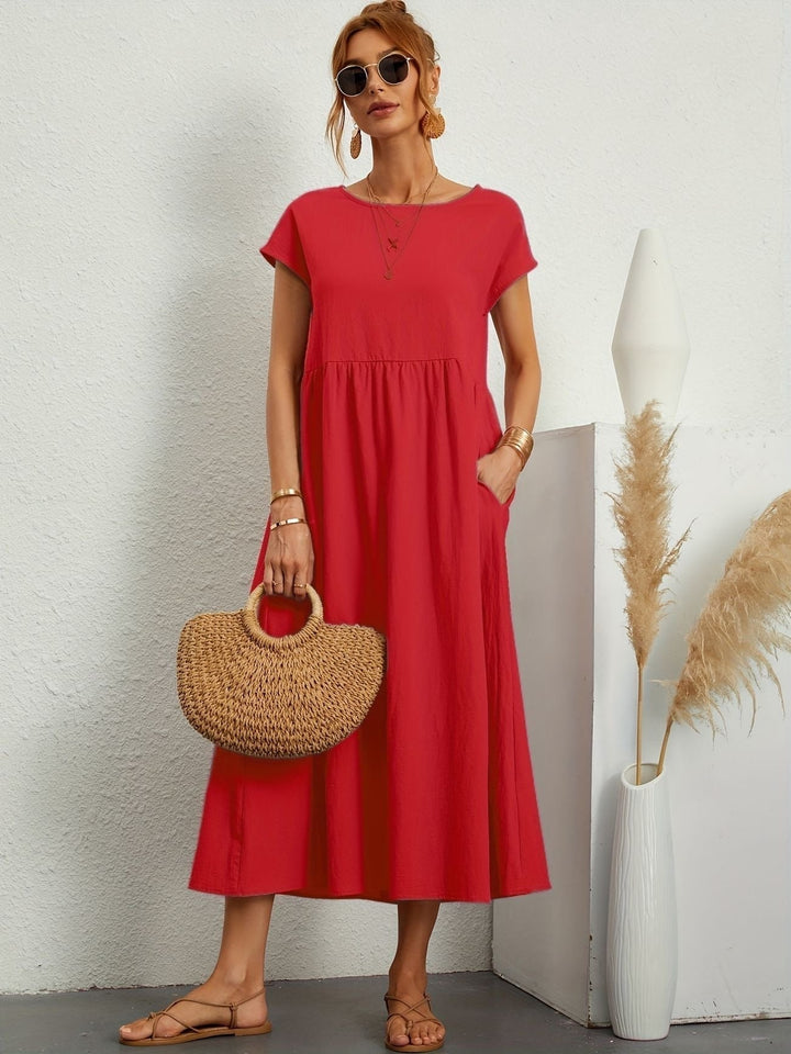 Donna con abito rosso estivo, borsa di paglia e sandali, posa accanto a vaso decorativo. Moda femminile casual, abbigliamento estivo elegante.