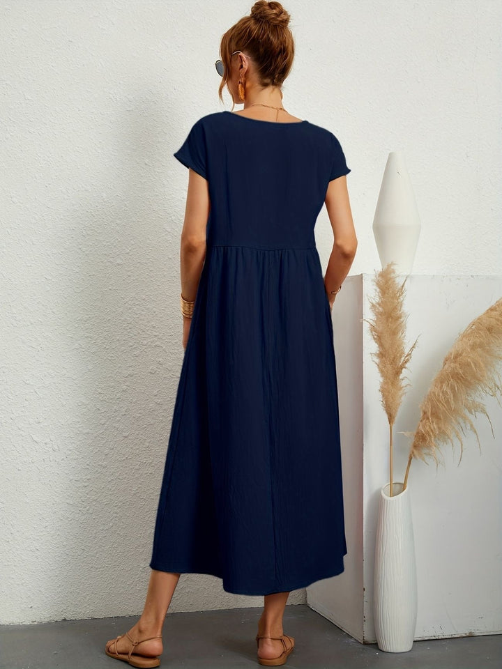 Donna con abito lungo blu, sandali beige, capelli raccolti, occhiali da sole, accanto a vaso bianco con pampas. Moda estiva elegante e casual.