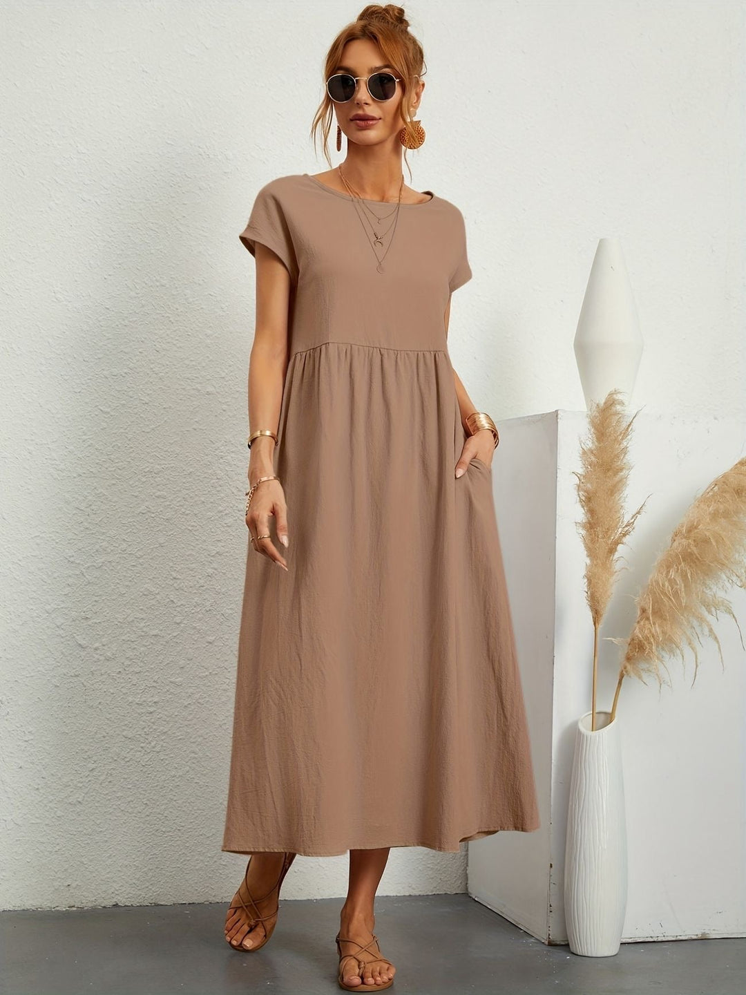 Donna con abito lungo marrone, occhiali da sole, sandali, accanto a vaso bianco con pampas. Moda estiva casual, abbigliamento femminile elegante.