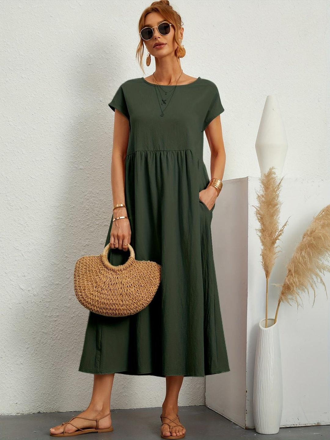 Donna con abito lungo verde, borsa di paglia e sandali, in posa accanto a vaso bianco con pampas. Moda estiva elegante e casual.