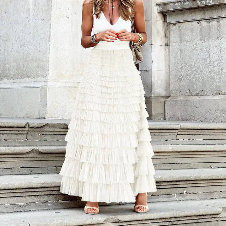 Donna con gonna lunga a balze bianche, top bianco senza maniche, sandali beige, su gradini di pietra. Moda estiva elegante, abbigliamento femminile.