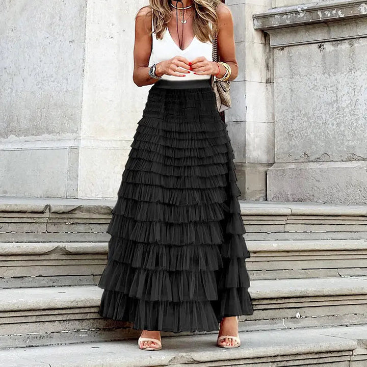 Donna con gonna lunga nera a balze, top bianco senza maniche, sandali beige, accessori colorati, su gradini di pietra. Moda elegante estiva.