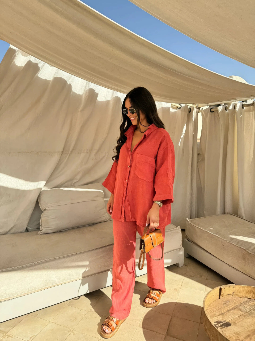 Donna con camicia e pantaloni rossi, occhiali da sole, borsa a tracolla marrone, sandali, in terrazza con tende bianche. Moda estiva casual.