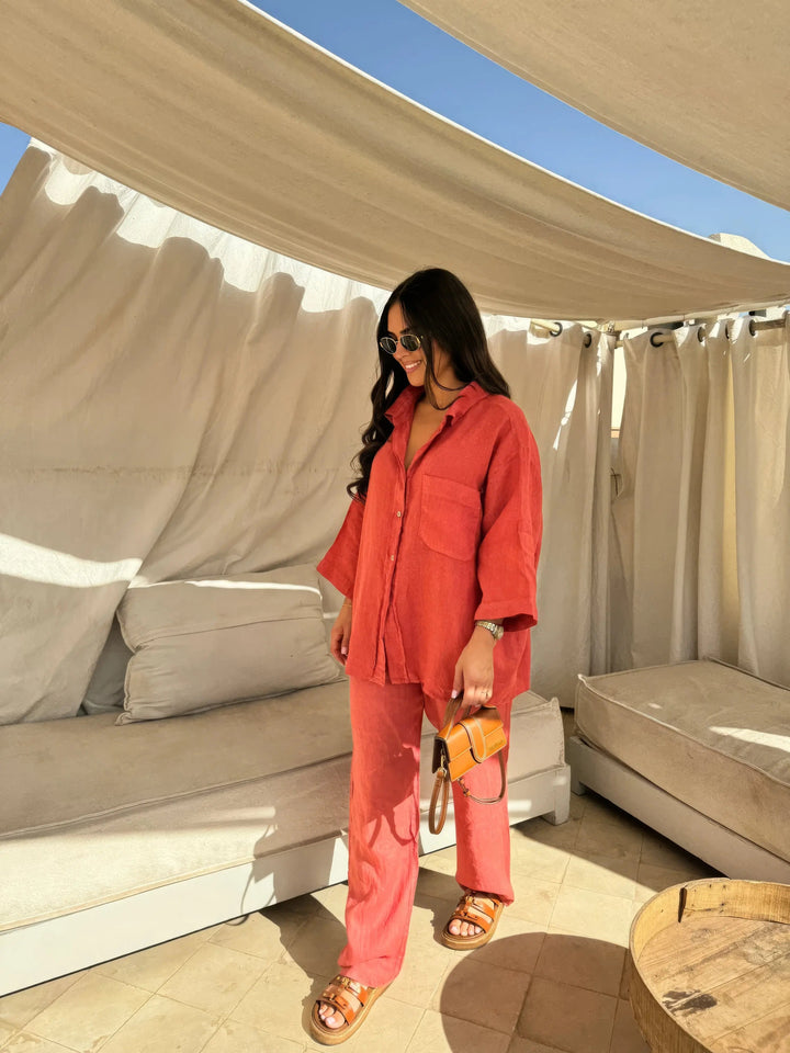 Donna con camicia e pantaloni rossi, occhiali da sole, borsa a tracolla marrone, sandali, in terrazza con tende bianche. Moda estiva casual.