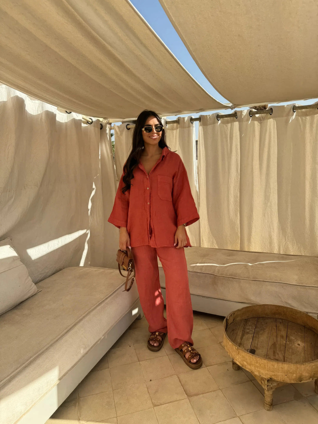 Donna con completo estivo arancione, camicia e pantaloni larghi, occhiali da sole, sandali marroni, borsa a tracolla, in terrazza con tende bianche.