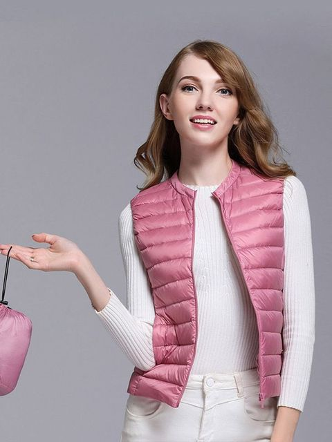 Giovane donna con gilet imbottito rosa, maglia bianca e pantaloni chiari. Moda autunnale, abbigliamento casual elegante. Sfondo grigio.