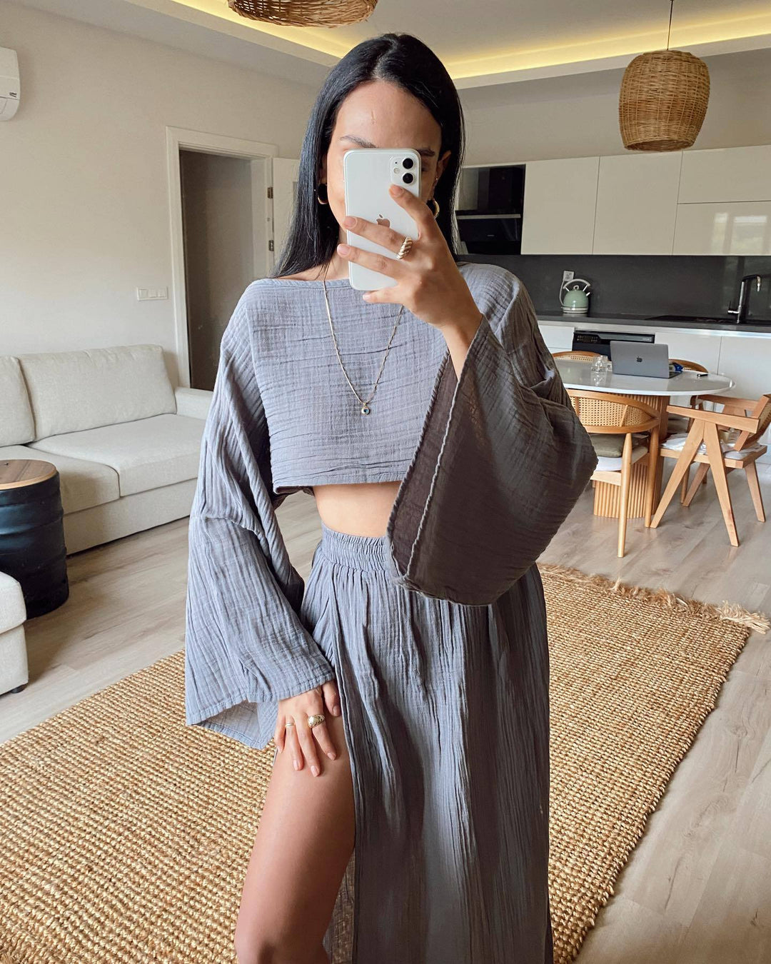 Donna in soggiorno moderno indossa abito coordinato grigio con top a maniche larghe e gonna lunga. Arredamento minimalista, tavolo in legno.