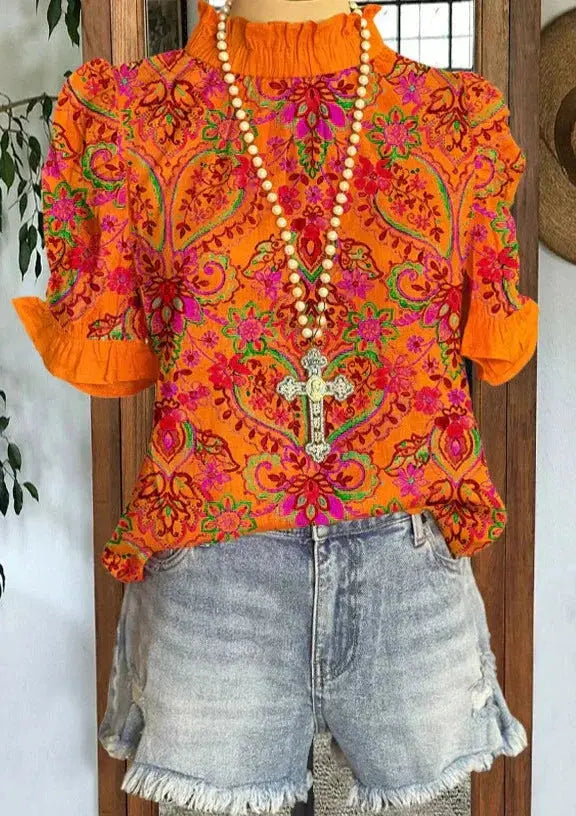 Blusa arancione con motivi floreali vivaci, collana con croce, abbinata a shorts di jeans sfrangiati. Moda estiva, abbigliamento casual donna.