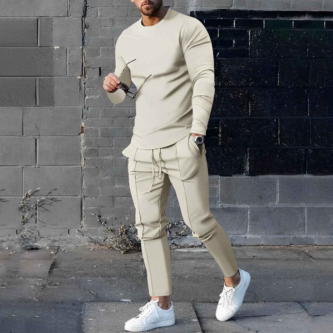 Uomo in tuta sportiva beige, maglia a maniche lunghe e pantaloni abbinati, indossa scarpe bianche, posa casual su sfondo urbano.