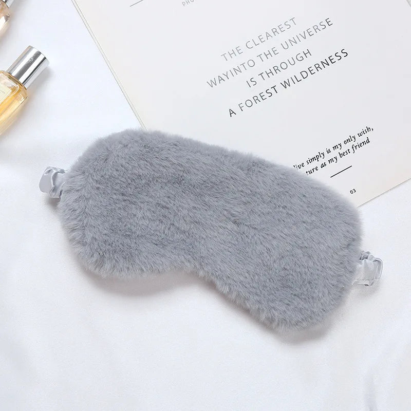 Mascherina per dormire in pelliccia sintetica grigia, morbida e confortevole, su sfondo bianco con libro e profumo. Accessorio relax e benessere.