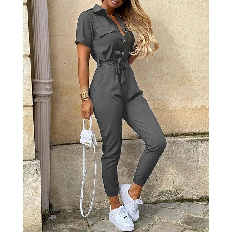 Tuta jumpsuit grigia da donna, maniche corte, con cintura in vita, abbinata a sneakers bianche e borsa a tracolla bianca. Moda casual elegante.
