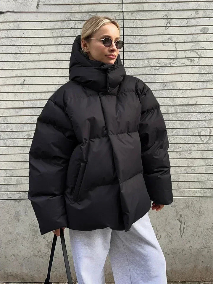Giacca invernale oversize nera, donna con occhiali da sole, pantaloni grigi, sfondo urbano. Moda streetwear, cappotto imbottito caldo.