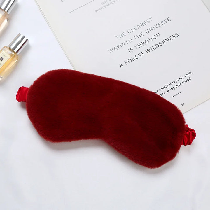 Mascherina per dormire in pelliccia sintetica rossa, elegante e morbida, su sfondo bianco con flaconi di profumo. Accessorio per il sonno di lusso.