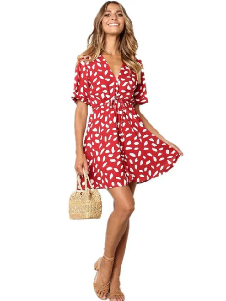 Abito estivo rosso a pois bianchi, maniche corte, scollo a V, indossato da donna sorridente. Accessori: borsa paglia, sandali beige.