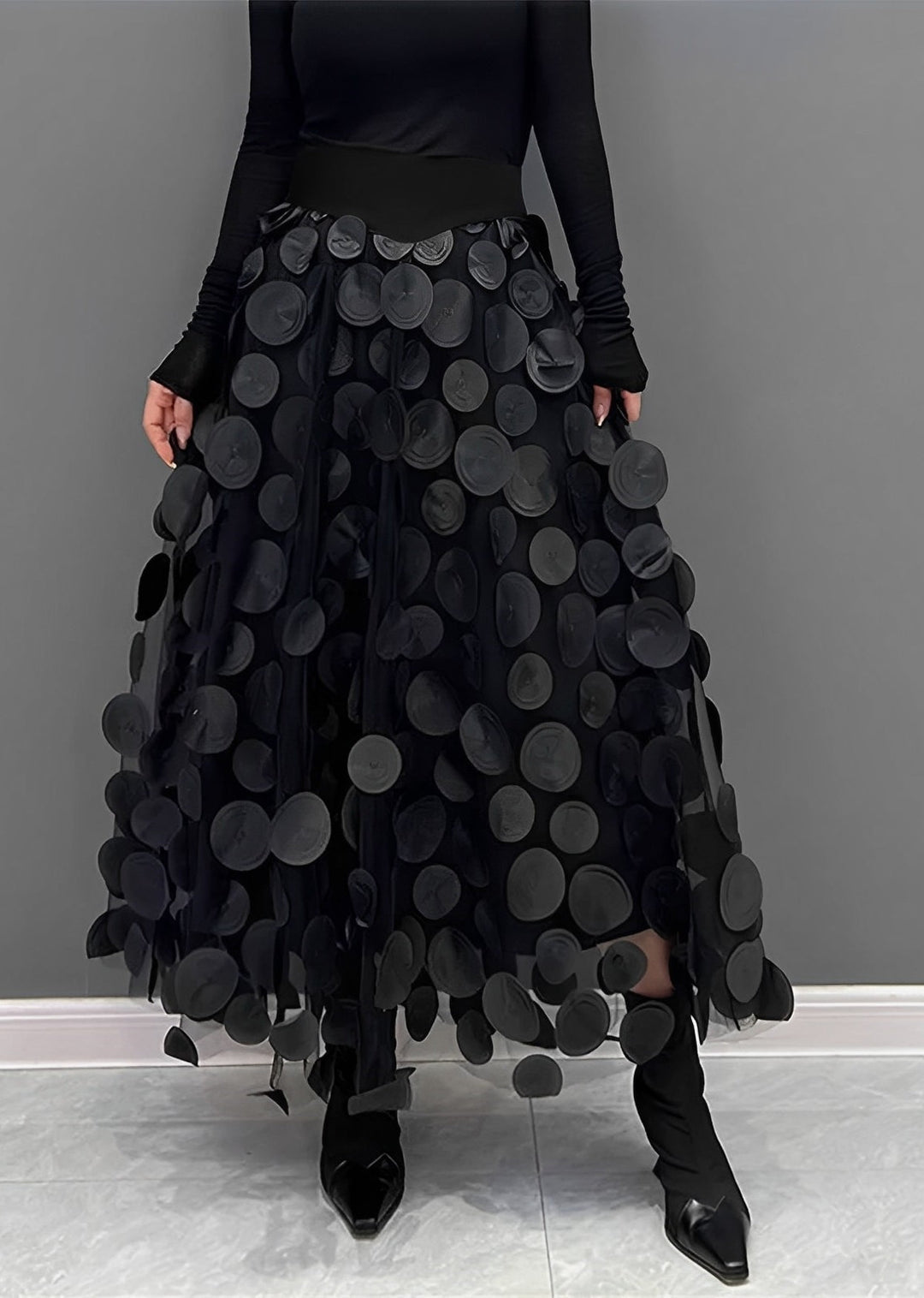 Gonna nera lunga con applicazioni circolari, indossata con stivali neri a punta. Design moderno ed elegante, perfetto per moda autunno/inverno.