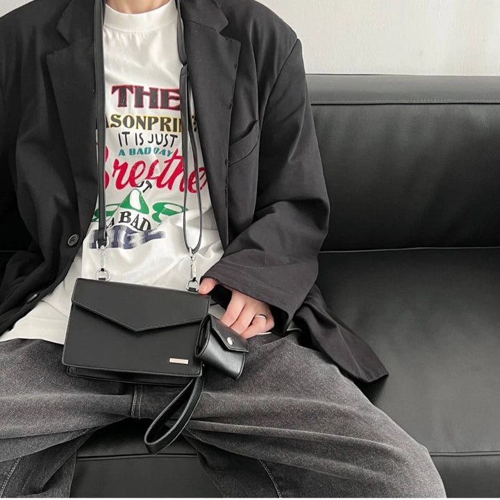 Giacca nera elegante su divano, borsa a tracolla nera in pelle, pantaloni grigi, maglietta bianca con stampa colorata. Moda casual uomo.