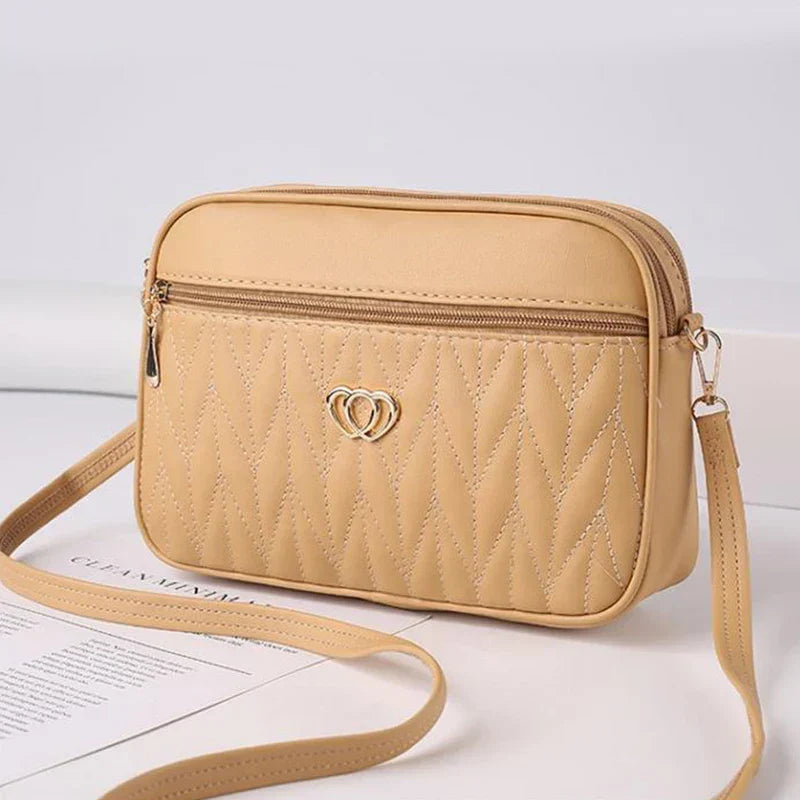Borsa a tracolla beige in pelle sintetica con motivo trapuntato e cerniera, decorata con logo a cuore dorato. Accessorio moda elegante e versatile.