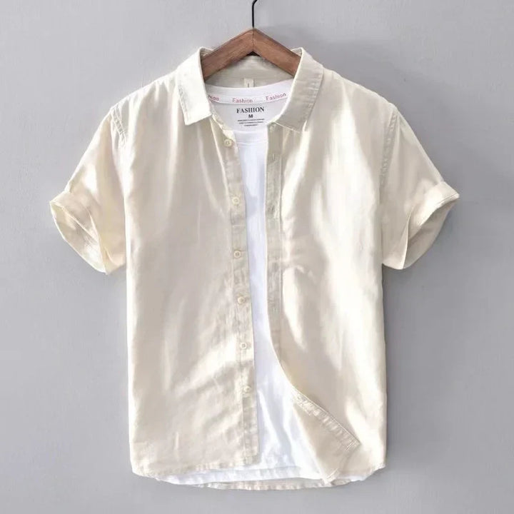 Camicia beige a maniche corte in cotone, appesa su gruccia di legno, sfondo grigio. Moda casual estiva, abbigliamento uomo elegante.