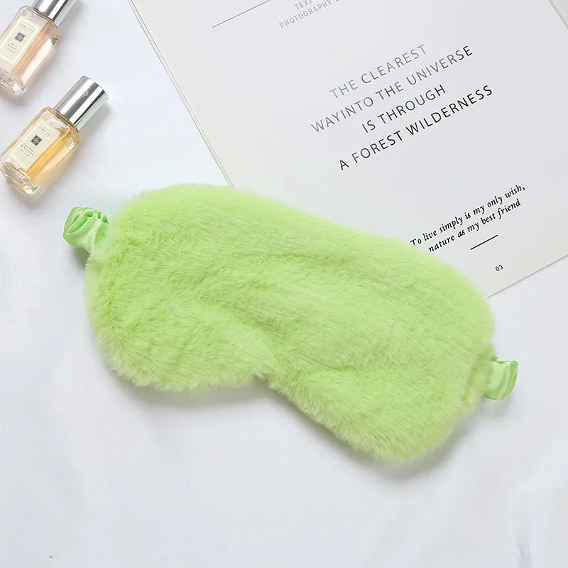 Mascherina per dormire verde in peluche su sfondo bianco, accanto a flaconi di profumo e libro. Accessorio relax, morbido e confortevole.
